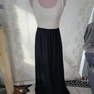 Elegant långklänning med spets och rosett - En elegant långklänning med en överdel i vit spets och en svart kjol. Klänningen har en stor rosett i ryggen och är ärmlös med en rund halsringning.