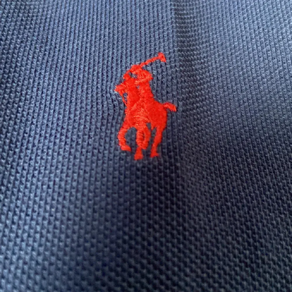 En mörkblå pikétröja från Polo Ralph Lauren med korta ärmar och en klassisk krage. Tröjan har en broderad röd logotyp på bröstet och knappar vid halsen. Är Xl men är som en L. T-shirts.