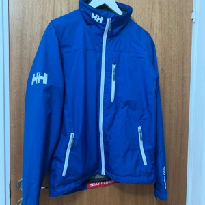 En blå helly Hansen jacka med bra kvalite  - Helly Hansen jacka med bra och skön matrerial. Den är emot regn och är perfekt för både regniga och blåsiga dagar.