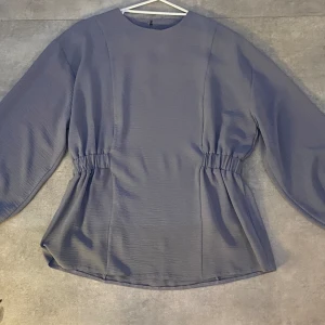 Blus- dovlilla - Långärmad blus med puff armar och knappar längst handled samt nacken. Fint resår längst midjan på höger och vänster sida. Den har ett tunt somrigt material och är endast använd en gång. Nytt skick utan prislapp.