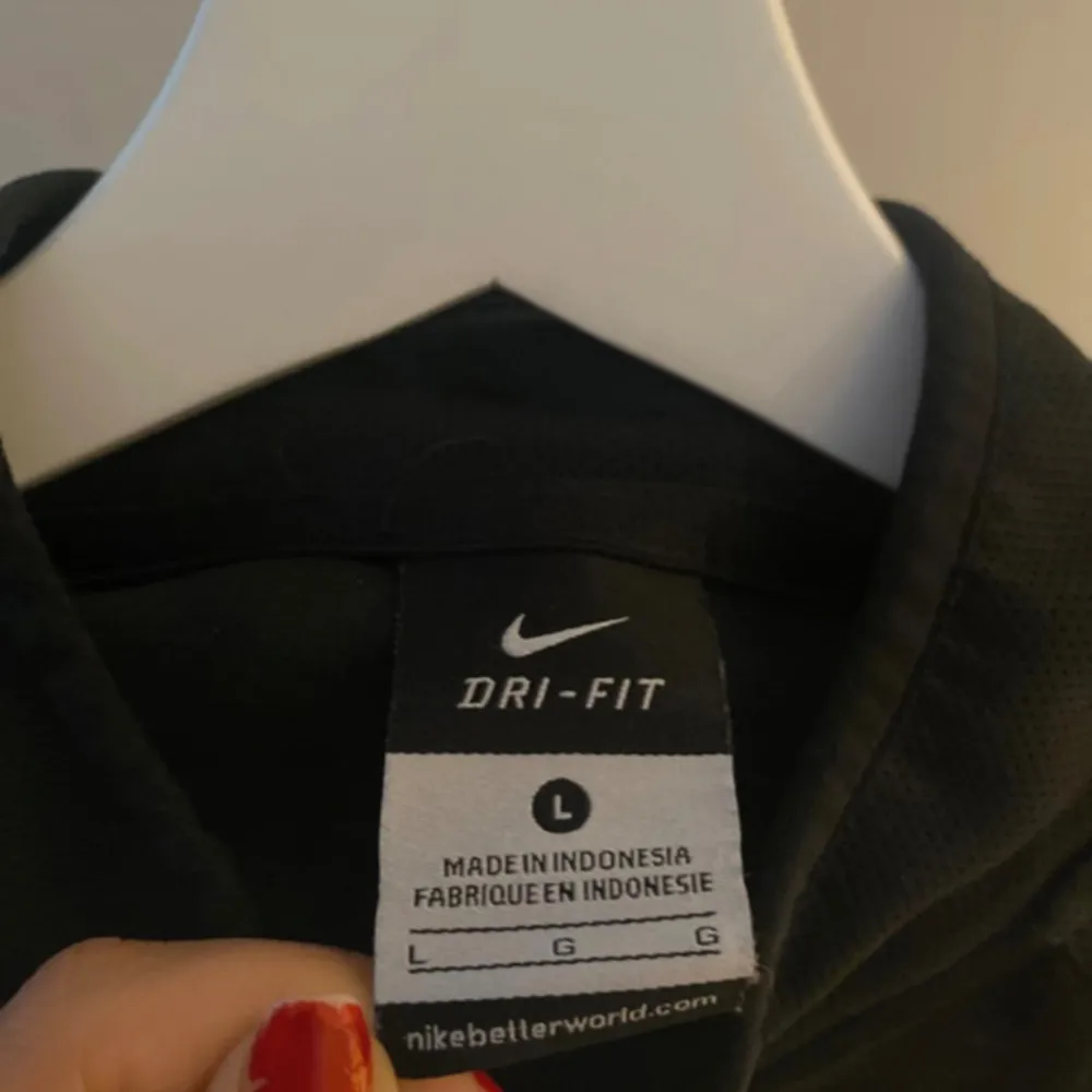 Svart jacka med dragkedja framtill och två fickor på sidorna. Logotypen är synlig på bröstet och på vänster ärm.. Sportswear.