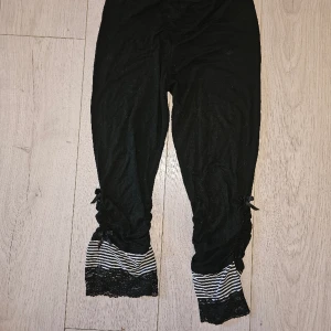 Svarta trekvartsleggings  - Beskrivning: Svarta piratbyxor med svartvitrandiga detaljer, spetsdetaljer samt rynkade detaljer på utsida ben. Märke: Storlek: 38/medium Skick: Mycket gott skick Material:   Nakenkatt finns i hemmet