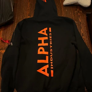 Alpha signature hoodie - Oanvänd, svart med orange tryck i storlek XL