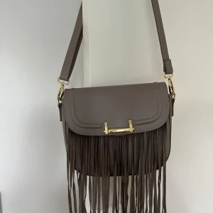 Väska Don Donna  - Säljer väska från Don Donna i mycket god skick, som ny, knappt använd. Beige färg. Imitationsläder, polyutetan/polyester.   Mått: Bredd 26 cm Höjd 20 cm (utan band)  Djup 8,5 cm Justerbara band som även går att ta av. 