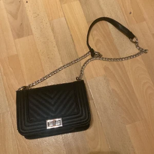 Svart axelremsväska  - En svart axelremsväska med silvrig kedja. Låsfunktion.