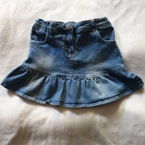 Blå jeanskjol med volang - En söt blå jeanskjol med volangdetalj längst ner. Kjolen har fickor fram och bak samt en knapp och dragkedja i midjan. Storlek 146