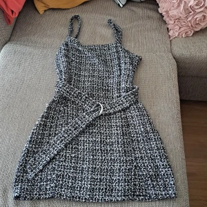 Svart och vit klänning med bälte - En elegant svart och vit klänning med ett matchande bälte. Klänningen har smala axelband och en rak skärning. Materialet är mönstrat i en tweed-liknande stil.