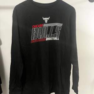 En svart långärmad tröja från NBA med Chicago Bulls logotyp och text i rött och vitt på framsidan. Tröjan har även text på ena ärmen.