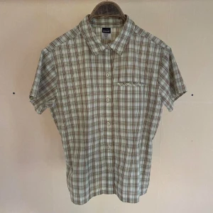 Rutig kortärmad skjorta från Patagonia - Patagonia. Vintage i fantastiskt skick. En bekväm, funktionell, rutig kortärmad skjorta från Patagonia. Skjortan har en klassisk krage, knäppning framtill och en bröstficka. Färgerna är en blandning av grönt, brunt och vitt.   -Material: 65% polyeste