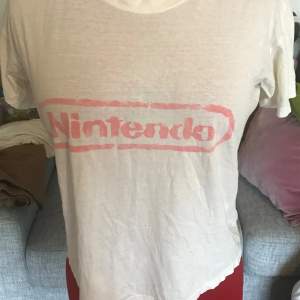 En vit t-shirt med korta ärmar och en röd Nintendo-logga tryckt på framsidan. T-shirten är i nyskick och har en normal passform.