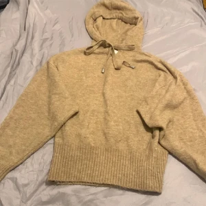 Beige stickad hoodie från H&M - En bekväm beige stickad hoodie från H&M. Hoodien har en justerbar dragsko vid huvan och ribbstickade muddar vid nederkanten. Perfekt för kyligare väder.