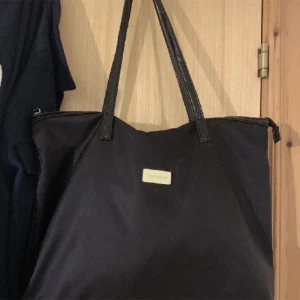 Svart handväska - En svart handväska med långa handtag och en liten rektangulär metallplatta på framsidan. Väskan har en enkel och stilren design.