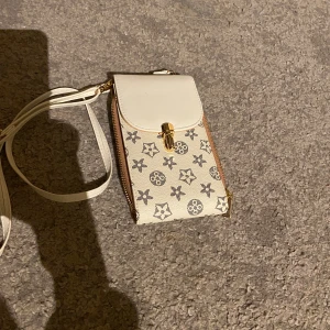 Vit axelväska med mönster - En liten vit axelväska med ett mönster av stjärnor och cirklar. Väskan har en dragkedja och en klaff med ett guldfärgat lås. Den har en justerbar axelrem.