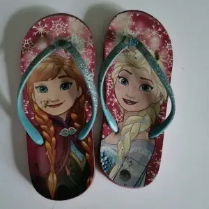 Ett par rosa sandaler med motiv från filmen Frost. Sandalerna har bilder av karaktärerna Anna och Elsa på sulan och glittriga remmar.