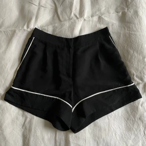 Svarta shorts med resår och vit detalj  - Svarta shorts från Monki. Resår baktill, knäppe framtill. Vit kant/detalj. Storlek 38. 