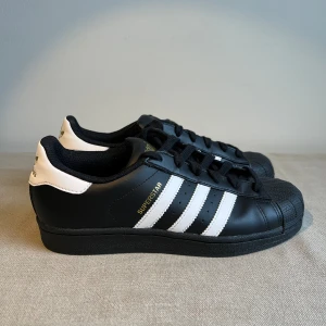 Adidas Superstar  - Helt oanvända Adidas Superstar i svart skinn, vita ränder och emblem i guld. Nypris  ca 1350 kr. Modellen är Unisex, herrstorlek FR 38/US 7.
