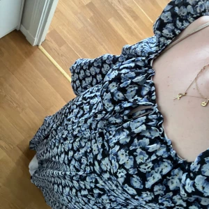 Blommig långklänning  - En blå och svart blommig långklänning med ballongärmar och knyte fram. Storlek 42, men passar om man har lite större M eller L. Går till golvet i längd på mig som är 171 cm. Aldrig använd och har prislappen kvar. Ordinarie pris 630kr