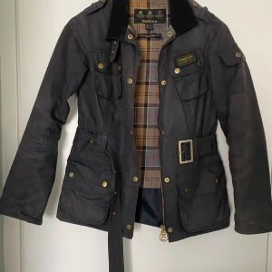 Vaxad Barbourjacka - Vår och höstjacka tunt fodrad. Fint använt skick. Slitage runt ärmar, krage och kanter. Du får med en oöppnad burk vax från Barbour. Normal i storlek