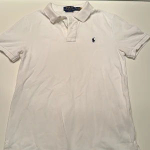 Vit slim fit t-shirt från Polo Ralph Lauren - Vit t-shirt från Polo Ralph Lauren i slim fit-modell. Klassisk pikékrage med knappar och broderad logga på bröstet. Tillverkad i bomull för en skön och luftig känsla. Perfekt för en clean och stilren look.