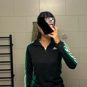 Svart Adidas träningströja XS - Aldrig använd med en prislapp. Svart långärmad Adidas träningströja med gröna och vita ränder längs ärmarna. Halv dragkedja framtill och klassisk Adidas-logga på bröstet. Tillverkad i lätt och funktionell polyester, perfekt för sport och aktivitet.