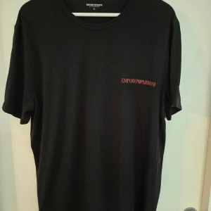 Svart t-shirt Emporio Armani XL - Svart t-shirt från Emporio Armani med diskret logga i rött på bröstet. Klassisk rund halsringning och korta ärmar. Tillverkad i mjuk bomull för en skön känsla och enkel stil.