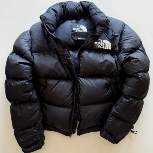 Svart pufferjacka från The North Face - Säljer en svart pufferjacka från The North Face med hög krage och dragkedja framtill. Jackan har en klassisk, puffig design och logga broderad på bröstet och insidan. Perfekt för kalla dagar och riktigt snygg streetstil.