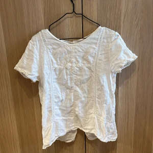 Vit blus med spetsdetaljer - Söt vit blus i bomull med korta ärmar och fina spetsdetaljer längs fram- och baksidan. Blusen har en fyrkantig spetsapplikation framtill och diskreta sömmar som ger en romantisk känsla. Perfekt till jeans eller kjol för en fräsch look. Är bara lite skrynklig men det är bara att stryka.
