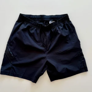 Svarta träningsshorts med snörning - Svarta shorts i syntetmaterial med elastisk midja och snörning framtill. De har en enkel och sportig design med två sidofickor och en loose passform, perfekta för träning eller chill dagar.