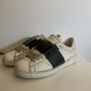 Valentino Garavani Open sneakers vit/svart - Säljer ett par Valentino Garavani Open sneakers i vitt skinn med bred svart läderdetalj över snörningen. Klassisk rund tå och platt sula, med grå snören och diskret logga på plösen. Snygg kontrast mellan färgerna och stilren design.