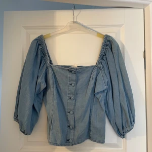 Ljusblå jeansblus från H&M - Supersnygg ljusblå jeansblus från H&M med puffiga långärmar och fyrkantig urringning. Blusen har knappar framtill och resår vid axlarna för en cool, avslappnad look. Perfekt att styla med höga jeans eller kjol för en trendig vibe.
