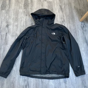Svart vind och regnjacka  från The North Face - Säljer en svart vind och regnjacka från The North Face med huva och dragkedja framtill. Jackan har två sidofickor med dragkedja och logga på bröstet. Perfekt för blåsiga och regniga dagar. Nypris 2299. Använd men väl omhändertagen. Pris går att förhandla. 