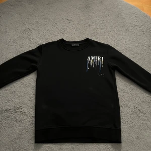  AMIRI sweatshirt - Svart sweatshirt från AMIRI med tryckt logga och färgstänk i blått, gult, vitt och brunt både fram och bak. Tröjan har rund hals, lång ärm och en clean, modern look. Perfekt för dig som gillar streetwear och vill sticka ut lite extra.
