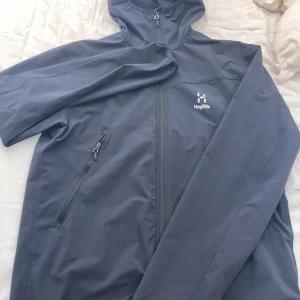 Svart vindjacka från Haglöfs, stl L - Säljer en stilren blå vindjacka från Haglöfs i storlek L, passar om du brukar bära M. Jackan har huva, hel dragkedja framtill och en praktisk ficka med dragkedja på bröstet. Perfekt för blåsiga dagar och outdoor-äventyr. Klassisk design med diskret Haglöfs-logga på bröstet.