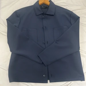 Mörkblå overshirt från ASOS Design - Stilren mörkblå overshirt från ASOS Design med klassisk krage, två bröstfickor och knäppning framtill. Jackan har en rak passform och är tillverkad i ett slätt polyestertyg som ger en clean look. Perfekt för lager-på-lager och enkel att matcha.