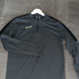 Svart Nike träningströja half zip - Svart långärmad träningströja från Nike med neongult swoosh på bröstet. Tröjan har en half zip-dragkedja framtill och är tillverkad i ett lätt, syntetiskt material. 