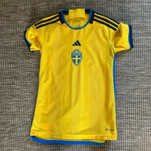 Svensk landslagströja Adidas gul XS - Gul fotbollströja från Adidas med blå detaljer och Sveriges landslagsemblem på bröstet. Tröjan har korta ärmar, blå ränder på axlarna och är tillverkad i ett lätt och ventilerande funktionsmaterial. Perfekt för dig som vill visa ditt stöd för Sverige på matchen.