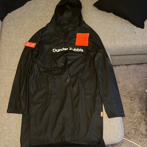 Svart regnjacka Dunder Zubbis Delux L - Svart regnkappa från odzxdelux med Dunder Zubbis-tryck på bröstet och en orange ficka som sticker ut. Kappan har huva, långa ärmar och stora fickor framtill. Materialet är syntetiskt och vattenavvisande, perfekt för regniga dagar.