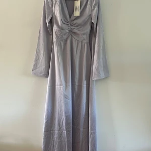 Ljuslila långklänning från Adoore - Supersnygg långklänning från Adoore i en laveldelnyans. Klänningen har v-ringning med rynkdetalj framtill och långa ärmar. Materialet har en lätt glansig finish som ger en lyxig känsla. Perfekt för dig som vill sticka ut med stilren elegans. Klänningen är ny med prislapp, endast provad