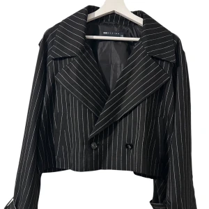 Svart croppad kavaj från ASOS Design - Snygg svart croppad kavaj med vita smala ränder från ASOS Design. Jackan har breda slag, dubbelknäppning och långa ärmar med knappar och spänndetaljer vid mudden. 