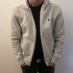 Grå hoodie från Polo Ralph Lauren - Snygg ljusgrå hoodie från Polo Ralph Lauren med dragkedja och huva. Tröjan har två fickor fram och klassisk Polo-logga broderad på bröstet. I mycket god skick och väl använd.