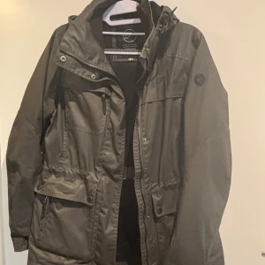 Mörk grön höstjack från Five Seasons - Snygg grön höstjacka från Five Seasons med huva, flera fickor med dragkedja och knappar samt justerbar midja. Jackan är vattenavvisande, vindtät och har meshfoder för extra komfort. Perfekt för dig som vill ha en funktionell och stilren jacka.