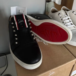 Christian Louboutin svarta sneakers 43,5 - Svarta sneakers från Christian Louboutin i skinn med ikonisk röd sula och silverfärgade nitar längs sidorna. Tungan har en svart, röd och vit detalj med logga. Snygg och edgy design som sticker ut.