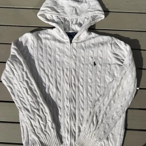 Vit kabelstickad hoodie från Ralph Lauren - Vit kabelstickad hoodie från Ralph Lauren i bomull. Tröjan har dragkedja framtill, huva och klassisk broderad logga på bröstet. Snygg ribbad mudd vid ärmslut och nederkant. Perfekt för en chill och stilren look.