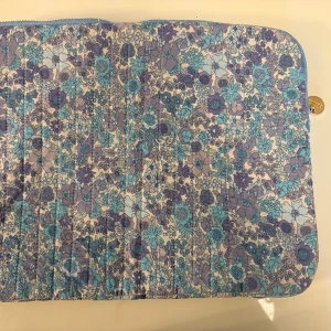 Blommig datorväska med dragkedja - Säljer en datorväska med blommigt mönster i blått, lila och beige. Väskan har quiltad yta och stängs med en guldfärgad dragkedja. Perfekt för att skydda din laptop och samtidigt ha en snygg accessoar.