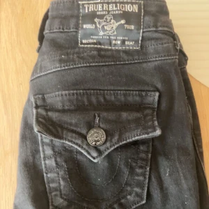 Svarta True Religion bootcut jeans - Säljer ett par svarta True Religion jeans, modell Becca Mid Rise Bootcut. Jeansen har snygga detaljerade bakfickor med knapp och klassisk logga. De är tillverkade i ett mjukt bomullsmaterial och har en bootcut passform som ger en schysst siluett.