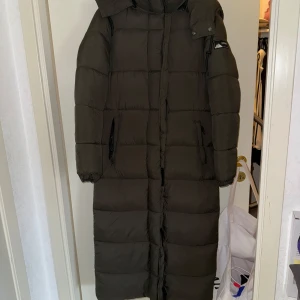 Lång mörkgrön pufferjacka Superdry - Säljer en lång och varm pufferjacka från Superdry i mörkgrönt med diskret rutmönster. Jackan har huva, dragkedja framtill och två fickor med dragkedja. Perfekt för kalla dagar och har en clean streetstyle-vibe.