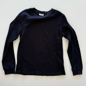 Svart långärmad tröja från Kappahl - En svart långärmad tröja från Kappahl med rund halsringning. Tröjan är ribbad och har en enkel, stilren design som passar perfekt till jeans eller under en jacka. Tillverkad i mjuk bomull för skön komfort.