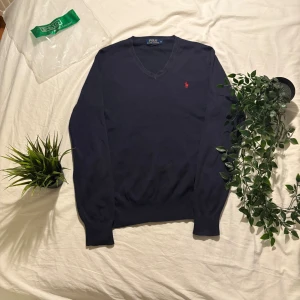 Mörkblå v-ringad tröja Polo Ralph Lauren - Klassisk mörkblå v-ringad tröja från Polo Ralph Lauren med röd broderad logga på bröstet. Långärmad modell i tunn, mjuk bomull som passar perfekt till lager-på-lager. Enkel och stilren design med ribbade muddar vid ärmslut och nederkant.