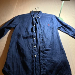 Mörkblå linneskjorta Polo Ralph Lauren XS - Snygg mörkblå skjorta från Polo Ralph Lauren i slim fit-modell. Tillverkad i linne med klassisk krage, vita knappar och det ikoniska röda broderade logot på bröstet. Perfekt för dig som gillar stilrena och tidlösa plagg.