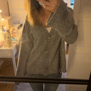 Grå stickad kofta med knappar - Mysig grå stickad kofta med v-ringning och stora knappar framtill. Koftan har en loose passform och långa ärmar med ribbade muddar. Perfekt att slänga på sig över ett linne eller t-shirt för en chill look.